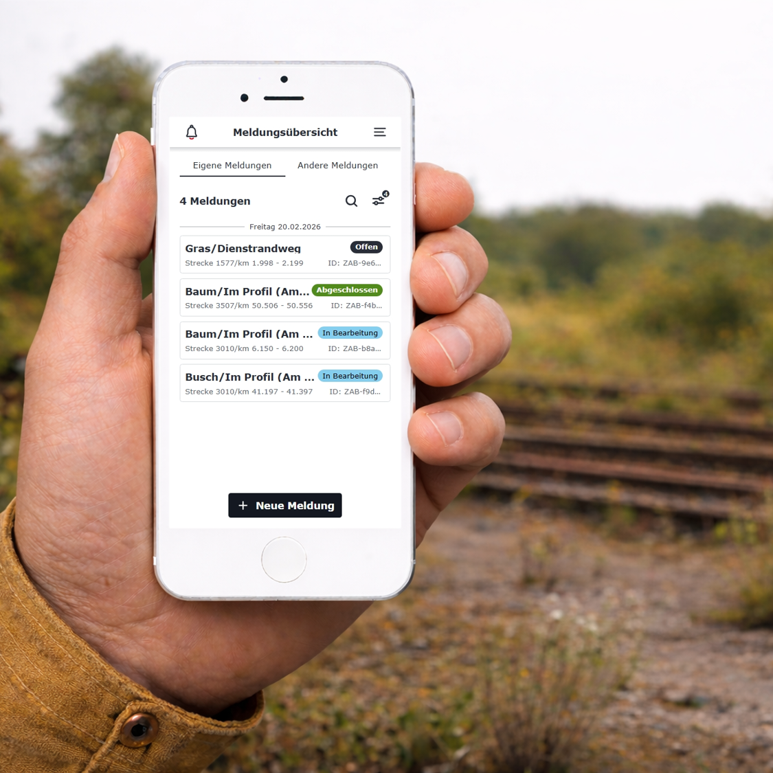 männliche Hand hält ein Smartphone in der linken Hand vor einem Hintergrund mit Gleisen, Büschen und Wald